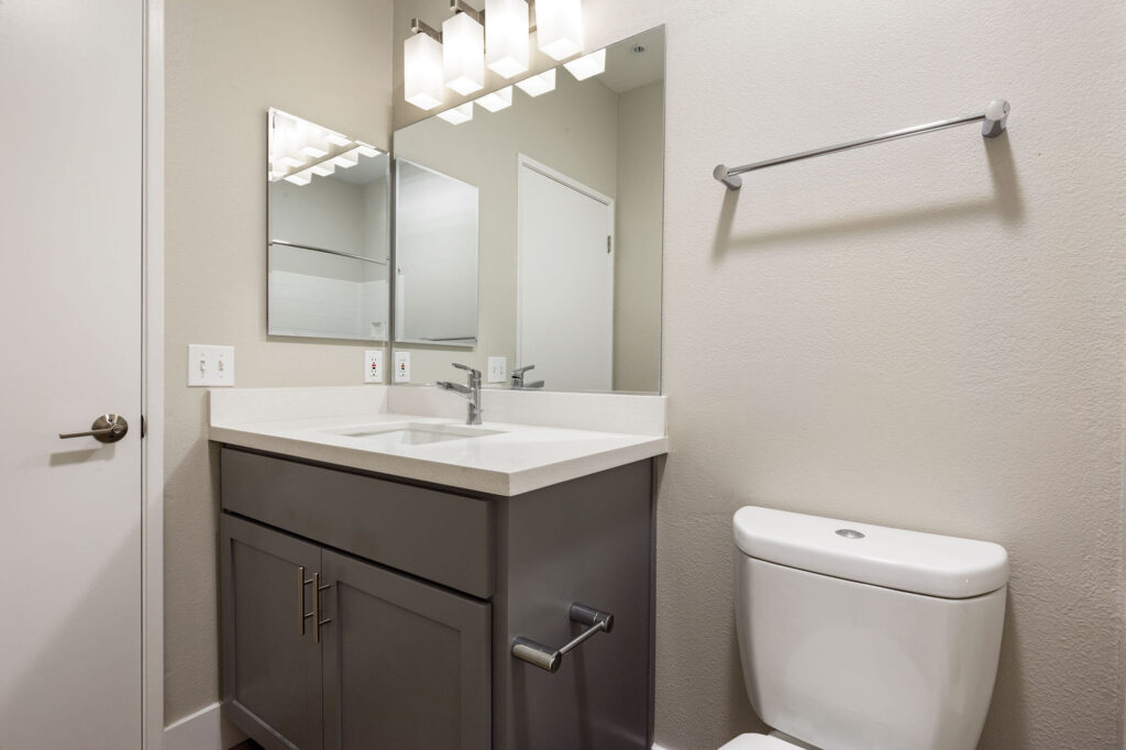 Bathroom with a grey vanity, white countertop, large mirror, and overhead lighting; located next to a standard toilet