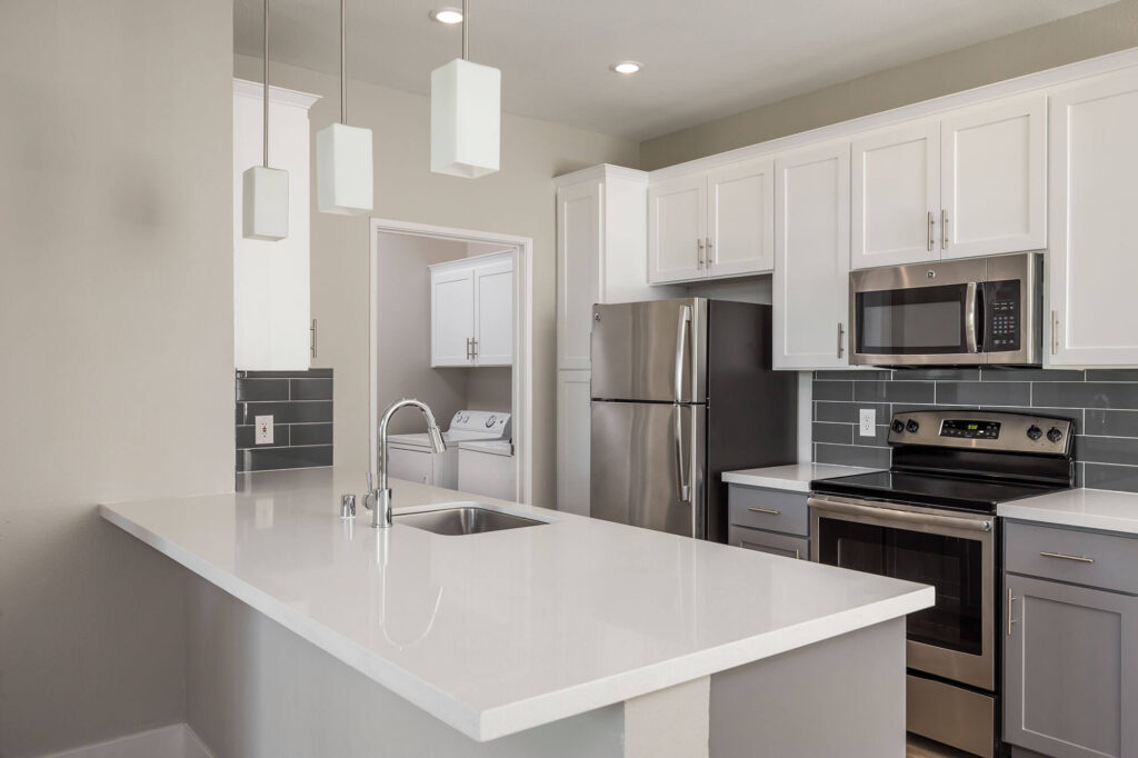 Galley-style kitchen featuring stainless steel appliances, grey cabinetry, white countertops, and a grey tile backsplash, with a view into the adjacent laundry room