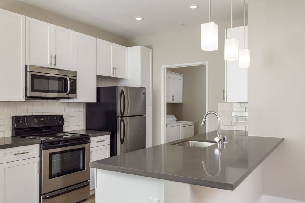 Galley-style kitchen featuring stainless steel appliances, white cabinetry, grey countertops, and white tile backsplash, with a view into the adjacent laundry room