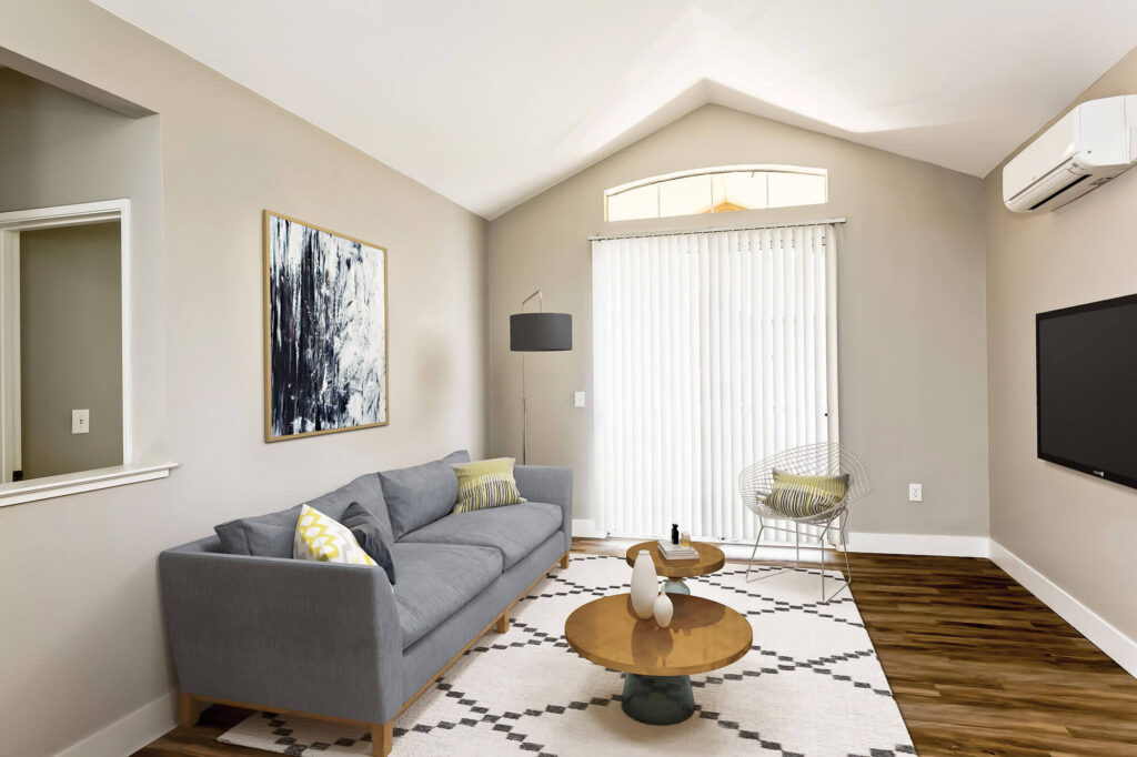 Living room with a large window and blinds, furnished with a sofa and armchair facing a television; wall-mounted air conditioning unit above the TV