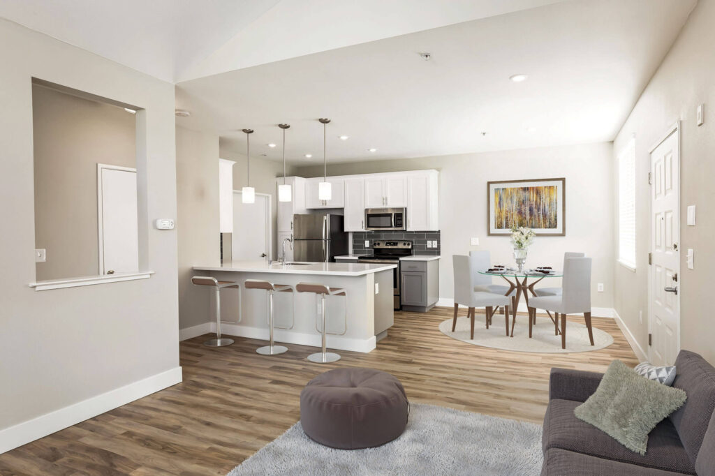 Kitchen with stainless steel appliances, gray cabinets, and white countertops, adjacent to a dining area with a table and four chairs near the front door; includes a view into the living room through a small wall cutout