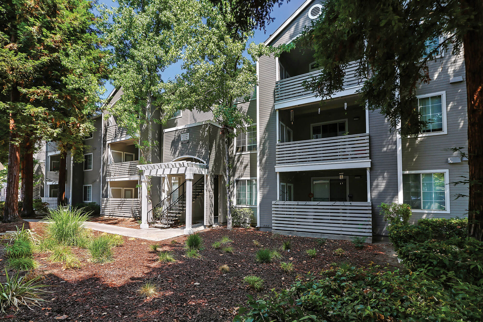 Grey apartment building exterior with covered patios and pathway through the landscaping