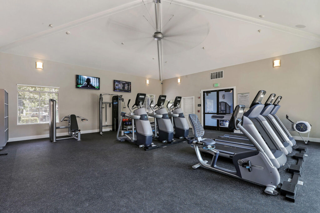 Indoor fitness center with cardio equipment, large overhead fan, and two televisions on the wall